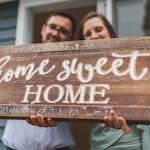 A joyful couple holding a 'home sweet home' sign in front of their new house.