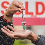 Close-up of hands exchanging house key with colorful SOLD sign in the background.