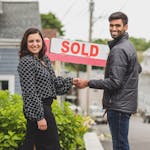 Two people shake hands in front of a sold sign, celebrating a real estate deal.