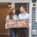 Young couple standing outside new home, holding 'home sweet home' sign, joyful and smiling.